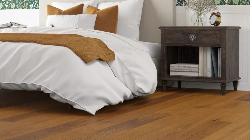 Warm neutral wood flooring beneath a white bed to advertise a 2026 floor trend: organic wood tones.