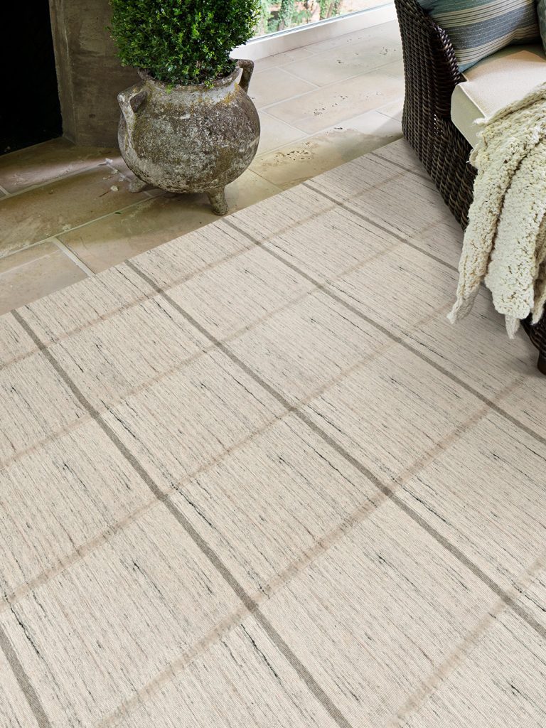 A beige and blue square patterned outdoor rug on a beige tile floor with a patio chair on one side and an outdoor fireplace visible on the side.
