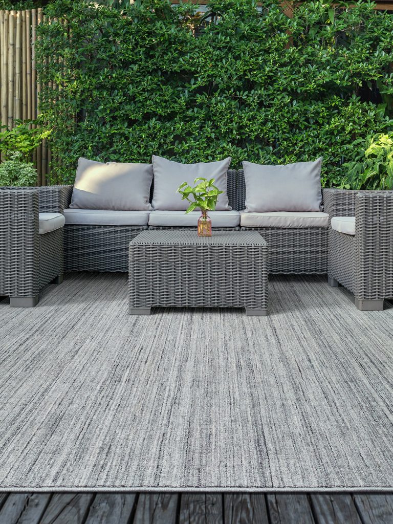 A gray textured outdoor rug on a gray wood patio with gray patio furniture and a green ivy colored wall in the background.