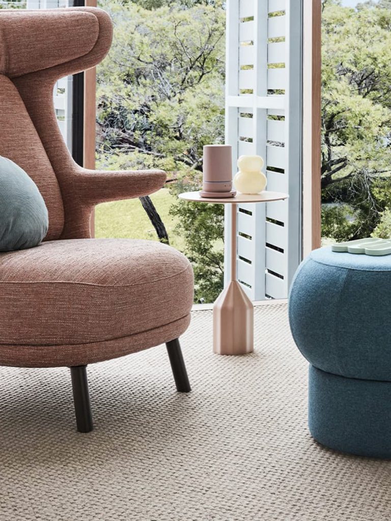 A pink armchair and blue ottoman on a beige carpet with the windows open in the background.