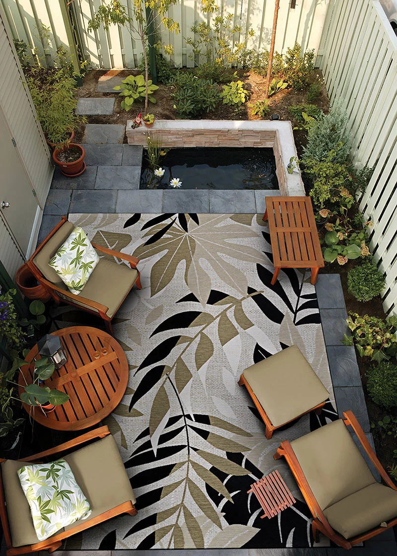 A brown, gray, and black leaf patterned indoor/outdoor rug on a stone patio surrounding by plants to advertise our Couristan rug and carpet sale.