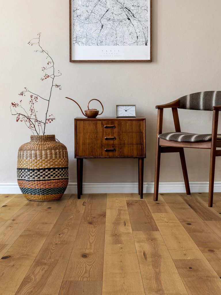 Shades of natural hardwood flooring with a vase, chair, and end table against a light colored wall.